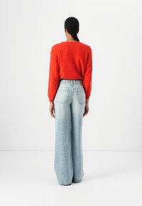 Rød teksturert genser med lange ermer kombinert med lyseblå vide jeans med baklommer og en synlig lærlapp i livet.