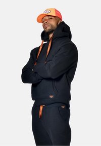 Un sweat à capuche noir et un pantalon avec des accents oranges et des cordons de serrage. Il dispose d'une poche kangourou et de patchs de logo. La casquette est d'un orange vif avec un dos en mesh.