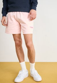 Lyserøde sweatshorts med sidelommer, elastisk talje med snor og broderet "WEEKEND" logo i lys pink. Båret med hvide sneakers og sokker.