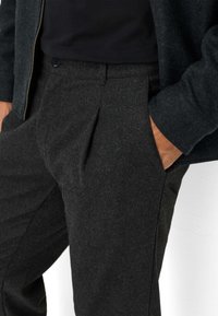 Donkergrijze, gestructureerde wollen broek met een geplooid voorpand, zijzakken en een aansluitend ontwerp, gedragen met een zwarte top en een donkere jas.