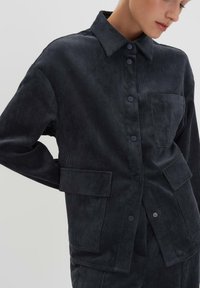 Donna che indossa una giacca in velluto a coste grigio scuro con bottoni a pressione e tre tasche applicate, in piedi davanti a uno sfondo chiaro e uniforme.