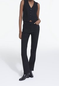 Gilet à rayures noires et pantalon assorti. Le gilet a un décolleté en V et une fermeture à boutons. Les pantalons sont ajustés avec une coupe slim. Bottes noires brillantes à la cheville.