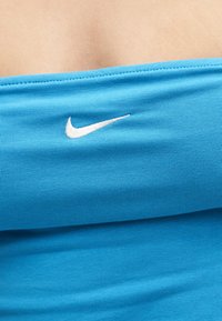 Μπλε ριμπάτο τοπ-σωλήνας με λευκό λογότυπο Nike στο κέντρο της πάνω πλευράς του στήθους, που αφήνει ακάλυφτο δέρμα πάνω από το ένδυμα.