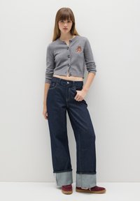 Cárdigan gris corto con bordado de oso, combinado con pantalones de mezclilla oscuros de pierna ancha que presentan un dobladillo de mezclilla clara. Zapatillas marrón y blancas.