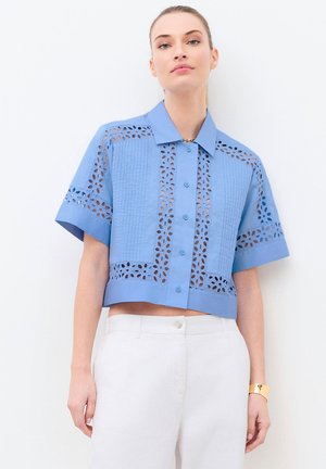 Vrouw draagt een lichtblauwe blouse met korte mouwen met bloemen uitgesneden patronen, gecombineerd met witte broek met hoge taille.