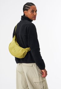 Grüner Nylon-Schultertasche mit geschwungener Form und runden Akzenten, getragen über einer schwarzen Fleecejacke und cremefarbenen Cargo-Hosen.