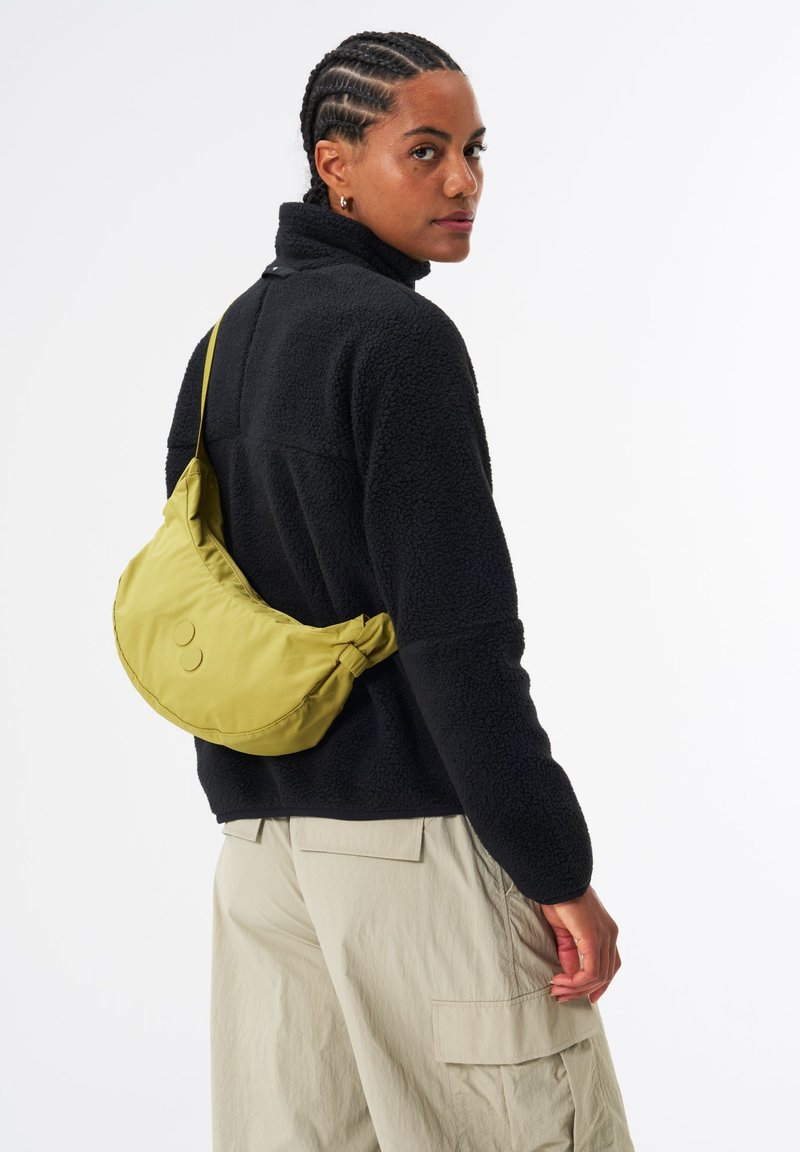 Grüner Nylon-Schultertasche mit geschwungener Form und runden Akzenten, getragen über einer schwarzen Fleecejacke und cremefarbenen Cargo-Hosen.