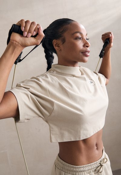 T-shirt de sport beige cropped avec un col montant, associé à un short à cordon de serrage assorti. Le sujet tient des bandes de résistance avec des poignées noires.