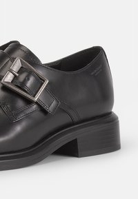 Scarpa in pelle nera con punta quadrata e una fibbia metallica argentata come accentuazione. Presenta un tacco basso impilato e una texture liscia.