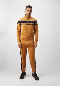 Welurowy dres w musztardowym kolorze z granatowymi paskami na ramionach i bokach, kurtka na zamek błyskawiczny, elastyczne mankiety i spodnie, zestawiony z czarnymi sneakersami.