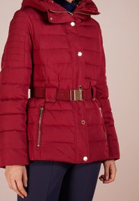 Punainen toppatakki, jossa vaakasuorat tikkaussomisteet, huppu, sivuvetoketjutaskut ja vyötärövyö. Sisältää kiiltävät metalliset yksityiskohdat.