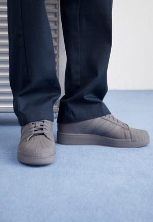 Person trägt dunkle Hose und graue Adidas Shell Toe Turnschuhe und steht auf hellblauem Teppich.