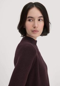 Frau mit kurzem dunklem Haar, die einen dunkelvioletten, gestrickten Pullover trägt und seitlich vor einem schlichten, hellen Hintergrund schaut.