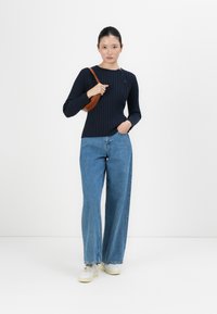 Frau steht mit einer Hand in der Jeans-Tasche und der anderen Hand hält eine braune Schultertasche, trägt einen dunklen Rippstrick-Pullover und Sneakers.