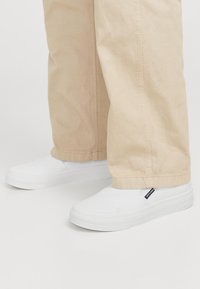 Fehér, sima vászon felületű slip-on cipők, lapos, gumitalppal és egy kis fekete címkével az oldalán, bézs nadrággal párosítva.