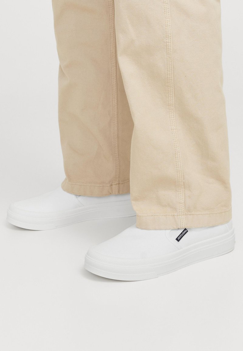 Fehér, sima vászon felületű slip-on cipők, lapos, gumitalppal és egy kis fekete címkével az oldalán, bézs nadrággal párosítva.