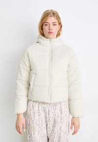 Giacca imbottita color crema con cappuccio aderente, zipper frontale e design imbottito. Indossata sopra pantaloni chiari a fantasia.