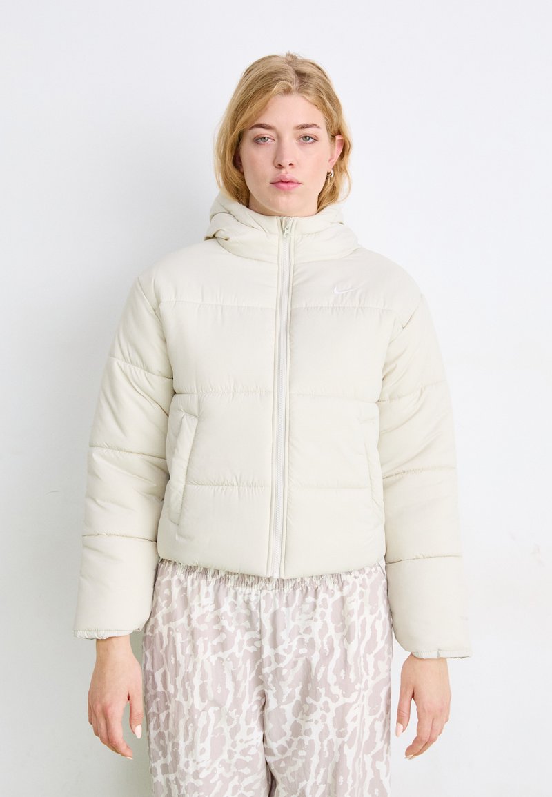 Giacca imbottita color crema con cappuccio aderente, zipper frontale e design imbottito. Indossata sopra pantaloni chiari a fantasia.