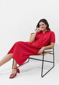 Rotes, kurzärmliges Kleid mit einem Reißverschluss und gerafftem Taillienbund, aus glattem Material gefertigt. Kombiniert mit roten Blockabsatzsandalen.