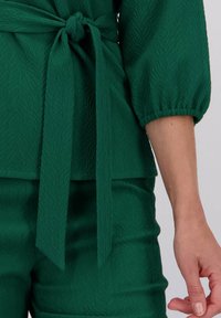 Groene gestructureerde stoffen outfit met een vastgebonden riem in de taille en driekwart pofmouwen, rustend op de arm van een persoon die naast hun lichaam staat.