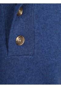 Blauwe gebreide polo met details van een gestructureerde stof, twee bruine knopen en een klein zakje. Het ontwerp is eenvoudig en functioneel.