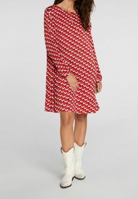 Robe rouge avec un motif géométrique blanc, à manches longues et col en goutte d'eau, fabriquée en tissu léger, assortie à des bottes de cowboy blanches.