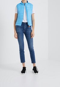 Veste sans manches matelassée bleu clair avec fermeture éclair sur le devant, portée sur une chemise blanche et associée à un jean bleu marine et des baskets noires.