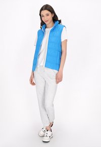 Gilet imbottito blu con colletto alto, chiusura zip, abbinato a una maglietta bianca e pantaloni di tuta grigi, indossato con sneakers bianche e nere.