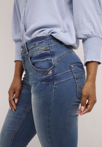 Visoko pasovi jeans s pošvikanim modrim opranjem, ki vključujejo šivano detajliranje, sprednje žepe in majhen logotip na strani.