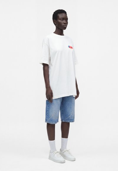Άτομο που στέκεται, φοράει λευκό oversized T-shirt με ένα μικρό πολύχρωμο γραφικό, μπλε τζιν σορτς με κείμενο, λευκές κάλτσες και λευκά αθλητικά παπούτσια.