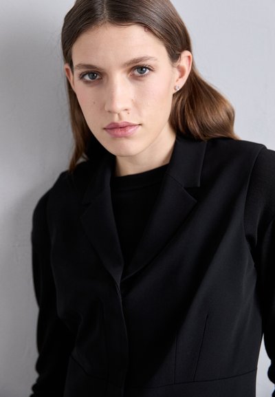 Melna pielāgota žakete ar asu atloku un strukturētu piegriezumu, ar tīrām līnijām un smalkiem šuvumiem, uzvilkta virs melna topa.