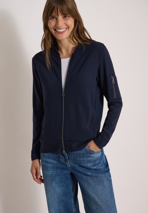 Femme souriante portant une veste zippée bleu marine avec une poche zippée sur la manche gauche et un jean bleu, debout devant un fond uni.