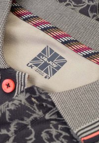 Donker patroon stof met subtiel bloemmotief, grijze textuur van de kraag, oranje knop, gestreepte afwerking en blauw label met de Britse vlag.