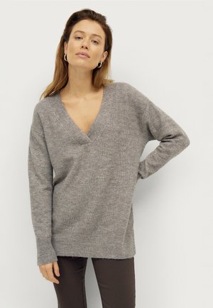 Grauer Strickpullover mit tiefem V-Ausschnitt, langen Ärmeln, gerippten Bündchen und lockerer Passform, kombiniert mit dunkelbraunen Hosen.