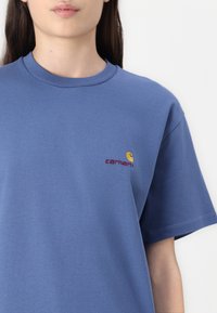 Blaues Baumwoll-T-Shirt mit Rundhalsausschnitt, kurzen Ärmeln und gesticktem Logo in Dunkelrot und Gelb auf der Brust. Glatte Textur.