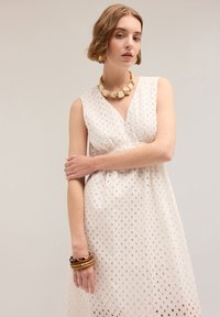 Rochie albă fără mâneci, cu decupaje texturate în formă de diamant, decolteu în V și pliuri delicate. Accesoriile includ un colier masiv bej și brățări suprapuse.