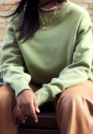 Person iført grøn sweatshirt med lagdelte guld- og perlesmykker, sidder med hænder sammenfoldet over brune bukser.