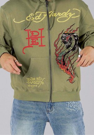 Sweat à capuche vert zippé avec des logos Ed Hardy rouges et jaunes et un motif serpent coloré sur le devant, assorti à un jean bleu avec logo assorti.