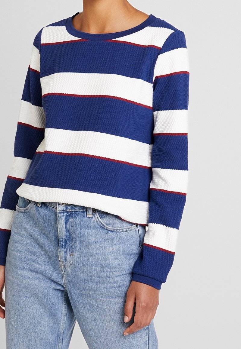 Femme portant un pull à manches longues rayé bleu et blanc avec de fines lignes rouges et un jean taille haute bleu clair.