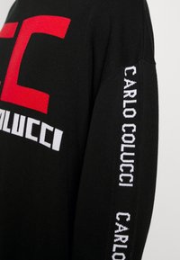 Czarny sweter wykonany na drutach z dużym czerwonym logo "C" oraz nazwą "CARLO COLUCCI" w białym kolorze na rękawie. Teksturowana tkanina z prążkowanym wykończeniem.