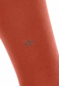 Primo piano di una calza di colore ruggine con texture, dotata di un sottile motivo a logo grigio, che enfatizza il tessuto e i dettagli del design.