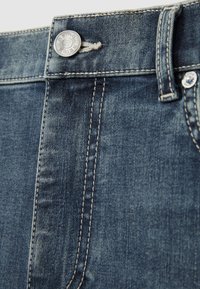 Detailný záber na pás modrých džínsov s strieborným gombíkom označeným "REISS" a viditeľnými prešívaniami.