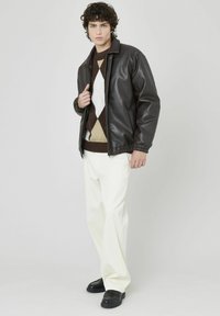 Veste en faux cuir marron avec un design à col, portée sur un pull argyle marron et crème. Associée à un pantalon blanc et des chaussures noires.