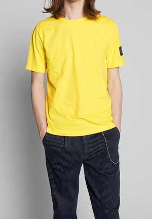 Żółta bawełniana koszulka z okrągłym dekoltem i krótkimi rękawami, zestawiona z granatowymi spodniami w prążki. Na rękawie znajduje się naszywka z logo.
