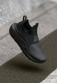 Schwarzer Sportschuh mit einer strukturierten synthetischen Oberseite, elastischem Riemen und runder Sohle. Besitzt Nike-Branding in einem dunkleren Farbton.