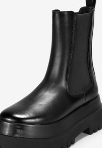 Botín negro de tobillo hecho de cuero brillante con paneles laterales elásticos, suela de goma negra gruesa y puntera redondeada, con un tirador en la parte trasera.