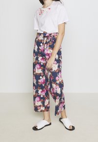 Nainen, joka on pukeutunut valkoiseen lyhythihaiseen paitaan, jossa on kukkakirjontaa, tummansinisiin kukkakuosisii leveisiin housuihin, joissa on vyö, ja valkoisiin liukuvaan sandaaleihin, seisoo sisätiloissa.