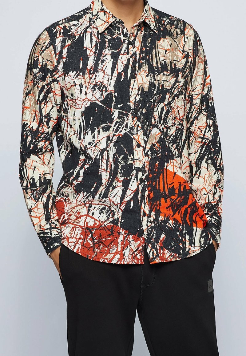 Chemise à manches longues boutonnée avec un motif abstrait de peintures éclaboussées en noir, blanc, orange et beige, associée à un pantalon noir.