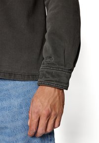 Manlig hand som vilar bredvid ljusblå denimjeans, iförd långärmad mörkgrå jacka med ett broderat krokodilmärke på ärmslutet.