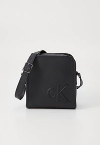 Svart läder axelväska med präglad "CK"-logotyp, fyrkantig form, dragkedjestängning och justerbar rem för mångsidighet. Mjuk textur.